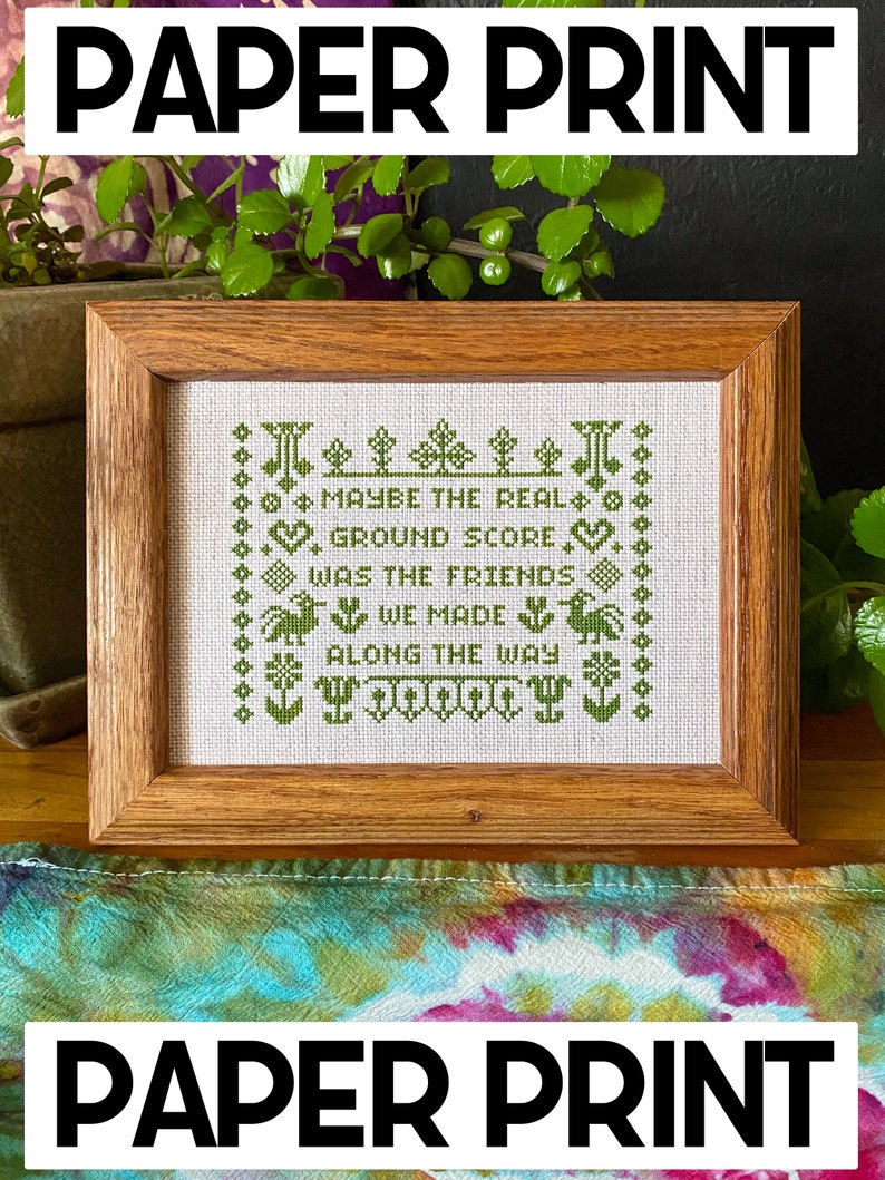 May include: A framed cross-stitch embroidery with green thread on white fabric. The embroidery reads "Maybe the real ground score was the friends we made along the way".