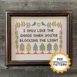 May include: A cross stitch pattern featuring a white background with the text "I only like the shade when you're blocking the light" surrounded by a green vine border. There are two blue birds and five green trees with orange trunks. The pattern is labelled "PDF Cross Stitch Pattern".