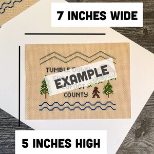 May include: A beige cross-stitch pattern with a brown bear, green trees, and blue waves. The pattern says "Tumble Down Sugar Creek County". The pattern is 7 inches wide and 5 inches high.