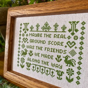 May include: A framed cross-stitch with green embroidery on a white background. The text reads "Maybe the real ground score was the friends we made along the way".