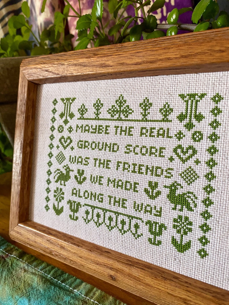 May include: A framed cross-stitch with green embroidery on a white background. The text reads "Maybe the real ground score was the friends we made along the way".