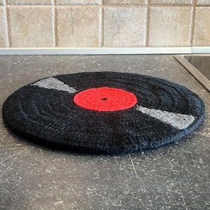 May include: A black, round, crocheted trivet designed to resemble a vinyl record. It features a red center and gray accent lines, adding a retro touch to kitchen decor. The trivet is placed on a dark countertop.