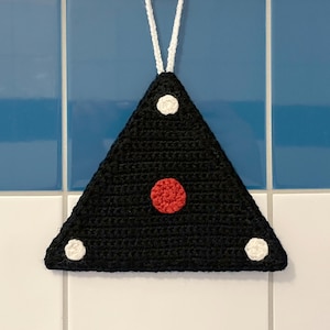 May include: A triangular black crochet potholder featuring a single red circular accent in the center and three smaller white circular accents at each corner, suspended by a white crocheted loop attached to a metal hook on a tiled wall.