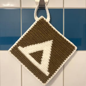 Può includere: Un presina all'uncinetto marrone e bianca con un design a triangolo bianco. La presina, di forma quadrata, ha un bordo bianco e un anello per appenderla. È appesa a un gancio di metallo su uno sfondo di piastrelle bianche e blu.