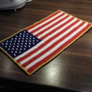 Könnte beinhalten: Eine gehäkelte amerikanische Flagge mit roten, weißen und blauen Streifen und einem blauen Feld mit weißen Sternen. Die Flagge ist mit einem gelben Besatz eingefasst.