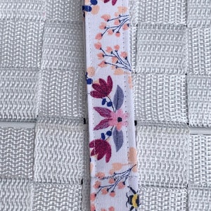 May include: A white fabric key chain with a floral and bee design. The key chain has a silver metal clasp.