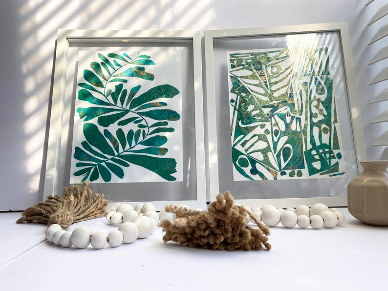 May include: Two framed botanical prints with green and gold leaf designs. Decorative elements include white beaded garlands and textured brown accents. A neutral-toned vase is also present, creating a cohesive aesthetic.