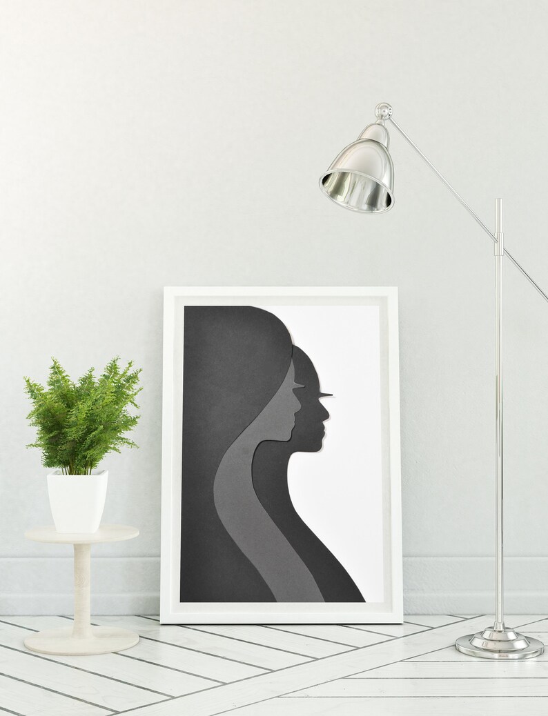 May include: A white framed print of a black and grey paper cut-out design of two women's profiles. The print is hanging on a white wall next to a silver floor lamp.