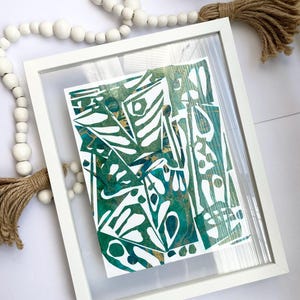 May include: Abstract art print in a white frame. The artwork features a mix of green, teal, and gold colours with organic shapes and patterns. The frame is rectangular and sits on a white surface, with a decorative beaded garland and tassel.