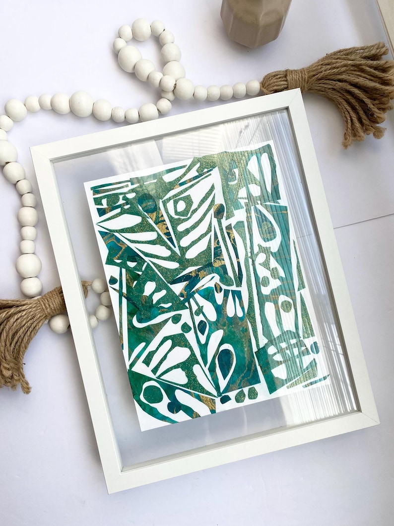 May include: Abstract art print in a white frame. The artwork features a mix of green, teal, and gold colours with organic shapes and patterns. The frame is rectangular and sits on a white surface, with a decorative beaded garland and tassel.