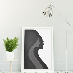 May include: A white framed print of a black and grey paper cut-out design of two women's profiles. The print is hanging on a white wall next to a silver floor lamp.