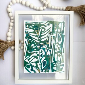 May include: Abstract art print in a white frame. The artwork features a green and gold design with white cut-out shapes. Decorative white wooden beads and a beige vase are also visible.
