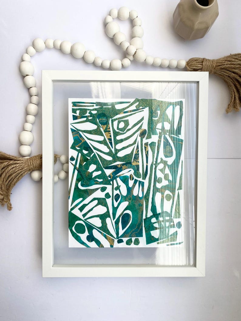 May include: Abstract art print in a white frame. The artwork features a green and gold design with white cut-out shapes. Decorative white wooden beads and a beige vase are also visible.