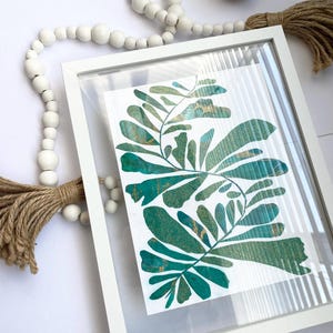 May include: A framed botanical print with teal and gold leaf designs. The artwork is in a white frame with a clear cover. White wooden beads and a jute tassel are also visible, adding to the decor.