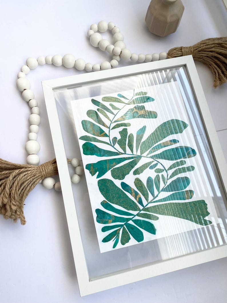 May include: A framed botanical print with teal and gold leaf designs. The artwork is in a white frame with a clear cover. White wooden beads and a jute tassel are also visible, adding to the decor.