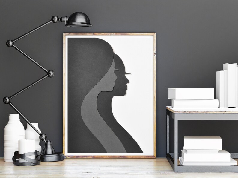 May include: A black and white framed print of a layered silhouette of two people's faces. The print is on a white background and is hanging on a grey wall.