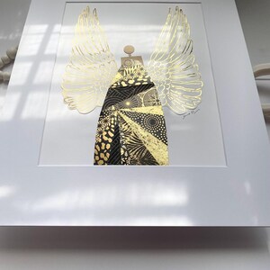May include: A framed print of a gold and black angel with wings. The angel is made of paper and has a geometric pattern. The print is signed "Jane Mann".