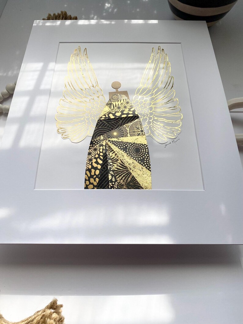 May include: A framed print of a gold and black angel with wings. The angel is made of paper and has a geometric pattern. The print is signed "Jane Mann".