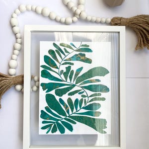 May include: A framed botanical print with teal and gold leaf patterns. The artwork is presented in a white frame, offering a contemporary look. The print's vivid colours and natural shapes bring a touch of nature-inspired decor to any space.