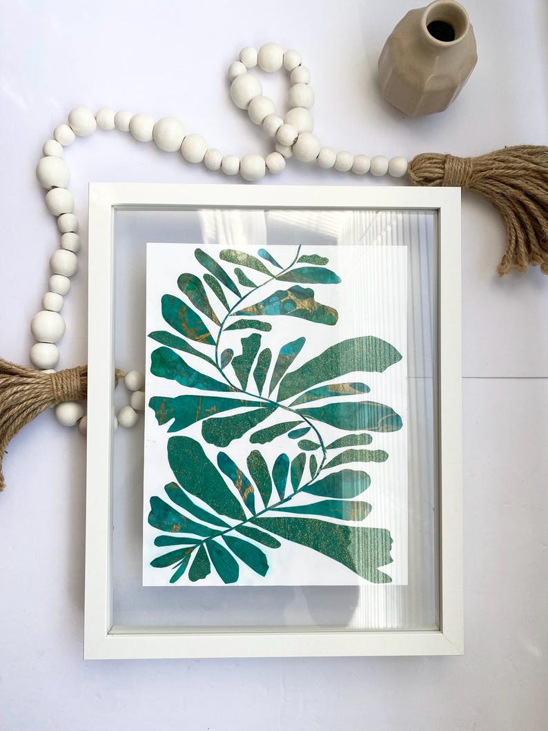May include: A framed botanical print with teal and gold leaf patterns. The artwork is presented in a white frame, offering a contemporary look. The print's vivid colours and natural shapes bring a touch of nature-inspired decor to any space.