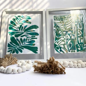May include: Two framed botanical prints with green and gold leaf designs. Decorative elements include white wooden bead garlands and textured brown accents. A neutral-toned vase is also present.