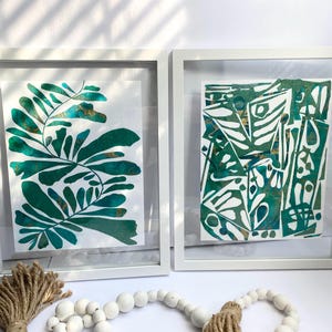 May include: Two framed abstract art prints. One features a green and gold leaf design, the other a geometric pattern in green and gold. Both are in white frames, creating a modern aesthetic. Decorative elements include a jute tassel and a beaded garland.