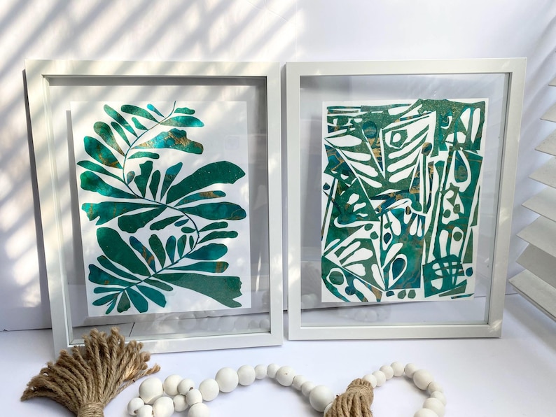 May include: Two framed abstract art prints. One features a green and gold leaf design, the other a geometric pattern in green and gold. Both are in white frames, creating a modern aesthetic. Decorative elements include a jute tassel and a beaded garland.