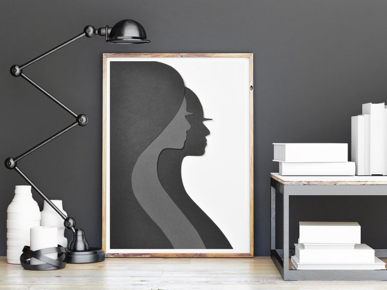 May include: A black and white framed print of a layered silhouette of two people's faces. The print is on a white background and is hanging on a grey wall.