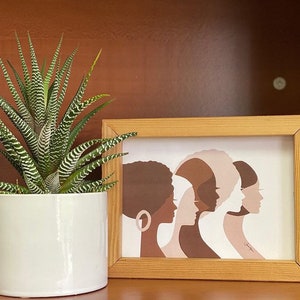 Peut inclure: Une impression encadr&eacute;e repr&eacute;sentant cinq profils de femmes dans des tons de marron, beige et blanc. L'&oelig;uvre est dans un cadre en bois clair, et une plante z&eacute;br&eacute;e en pot avec des feuilles ray&eacute;es vertes et blanches est &agrave; gauche.