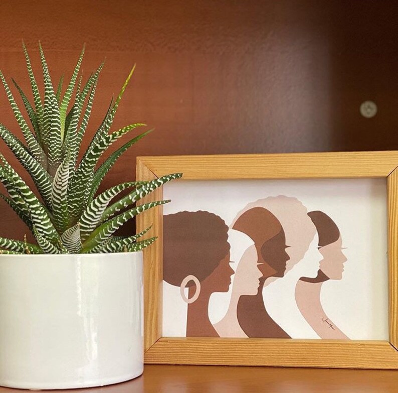 Peut inclure: Une impression encadr&eacute;e repr&eacute;sentant cinq profils de femmes dans des tons de marron, beige et blanc. L'&oelig;uvre est dans un cadre en bois clair, et une plante z&eacute;br&eacute;e en pot avec des feuilles ray&eacute;es vertes et blanches est &agrave; gauche.