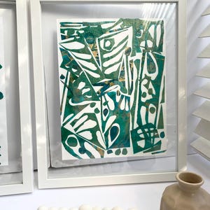 May include: Abstract art print in a white frame. The artwork features a mix of green, white, and gold shapes. The framed art is displayed on a white surface, with a decorative vase and wooden bead garland in the foreground.