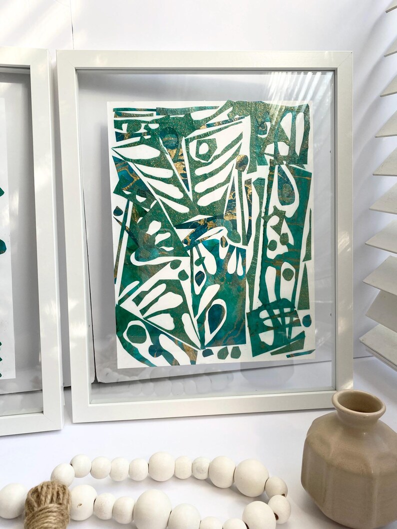 May include: Abstract art print in a white frame. The artwork features a mix of green, white, and gold shapes. The framed art is displayed on a white surface, with a decorative vase and wooden bead garland in the foreground.
