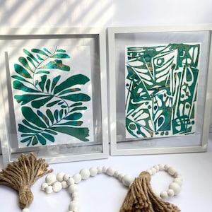May include: Two framed abstract art prints in white frames. One shows green leaf designs, the other a geometric pattern in green and white. A decorative tassel and beaded garland are in the foreground.