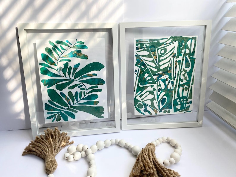 May include: Two framed abstract art prints in white frames. One shows green leaf designs, the other a geometric pattern in green and white. A decorative tassel and beaded garland are in the foreground.