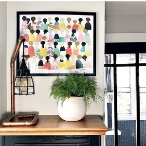 May include: A framed print of a colorful abstract art piece featuring a group of people in various shades of pink, yellow, green, and blue. The print is hanging on a white wall above a wooden table with a copper pipe lamp and a potted fern.