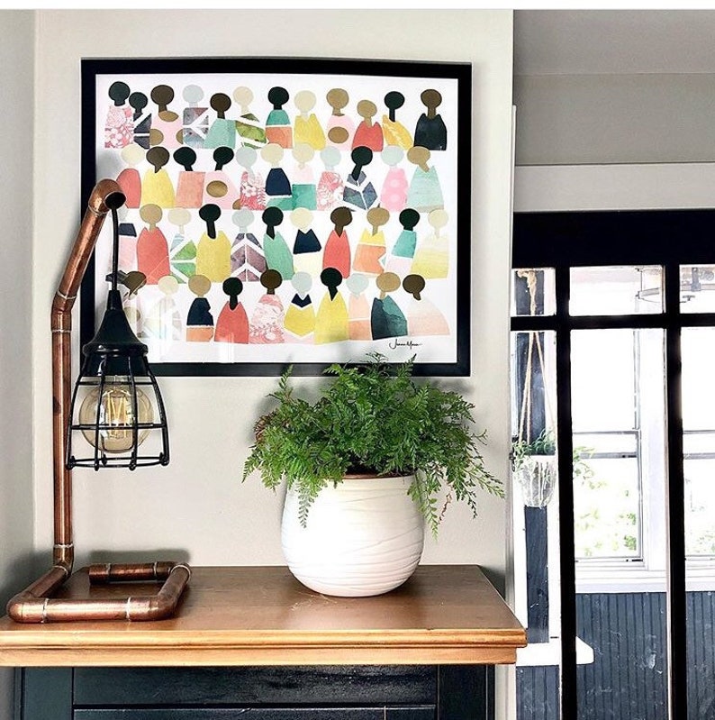 May include: A framed print of a colorful abstract art piece featuring a group of people in various shades of pink, yellow, green, and blue. The print is hanging on a white wall above a wooden table with a copper pipe lamp and a potted fern.