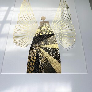 May include: A gold and black geometric angel with wings, the angel is made of paper and has a textured surface. The artwork is signed "Jane M."
