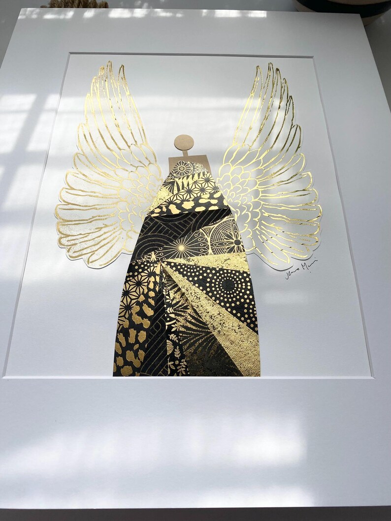 May include: A gold and black geometric angel with wings, the angel is made of paper and has a textured surface. The artwork is signed "Jane M."