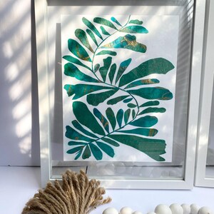 May include: A framed botanical art print with green and gold leaf designs. The artwork is in a white frame, with a bundle of twine and a strand of white wooden beads in the foreground. The art print is on a white background.