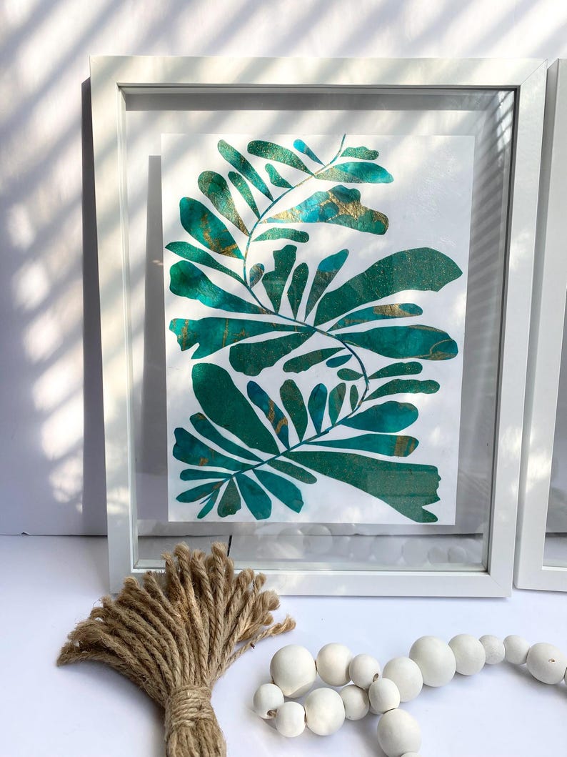 May include: A framed botanical art print with green and gold leaf designs. The artwork is in a white frame, with a bundle of twine and a strand of white wooden beads in the foreground. The art print is on a white background.