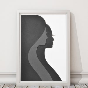 May include: A framed black and white paper art print of two overlapping silhouettes of people's profiles. The artwork is in a white frame and is on a wooden floor.