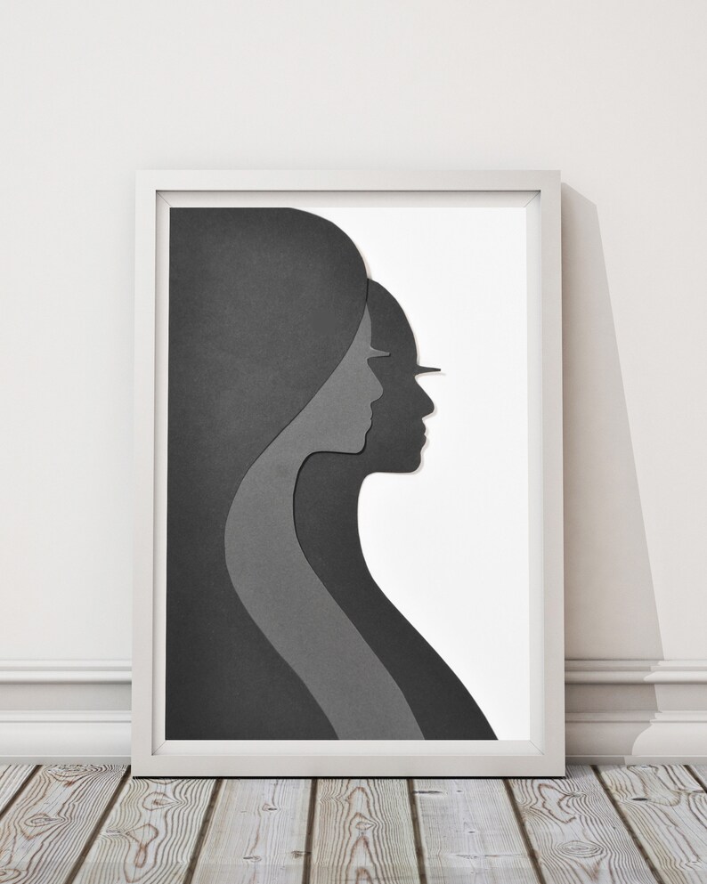 May include: A framed black and white paper art print of two overlapping silhouettes of people's profiles. The artwork is in a white frame and is on a wooden floor.