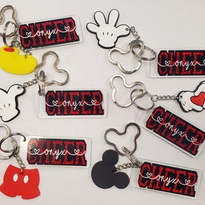 May include: Seven keychains with red and black glitter backgrounds. Each keychain features a different charm, including Mickey Mouse ears, a hand, and a heart. The keychains also have the text "CHEER onyx" on them.