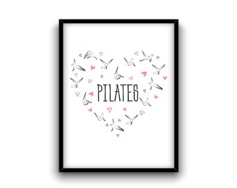 May include: A black framed print with a white background. The print features a heart shape made of black stick figures in various yoga poses. The word "PILATES" is written in black text inside the heart. Pink hearts are scattered around the figures.