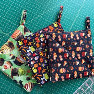May include: Three colorful, square pot holders with hanging loops. One is green with cupcakes, another is black with Halloween icons, and the third is dark blue with coffee and pastries. The pot holders are made of fabric.