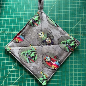 May include: A gray quilted pot holder with a hanging loop. The fabric features a repeating pattern of Jack Skellington and Sally from the Nightmare Before Christmas.