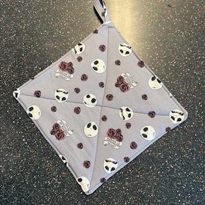 May include: A gray and white cotton pot holder with a repeating pattern of Jack Skellington's face and red roses. The text "Master of Fright" is printed on the pot holder.