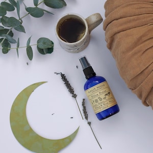 May include: A blue bottle of pillow and room mist with the text "Salix Moon Apothecary" and "Pillow Potion" on the label. The bottle is sitting on a white surface with a green crescent moon, lavender sprigs, and a brown ceramic mug with tea.