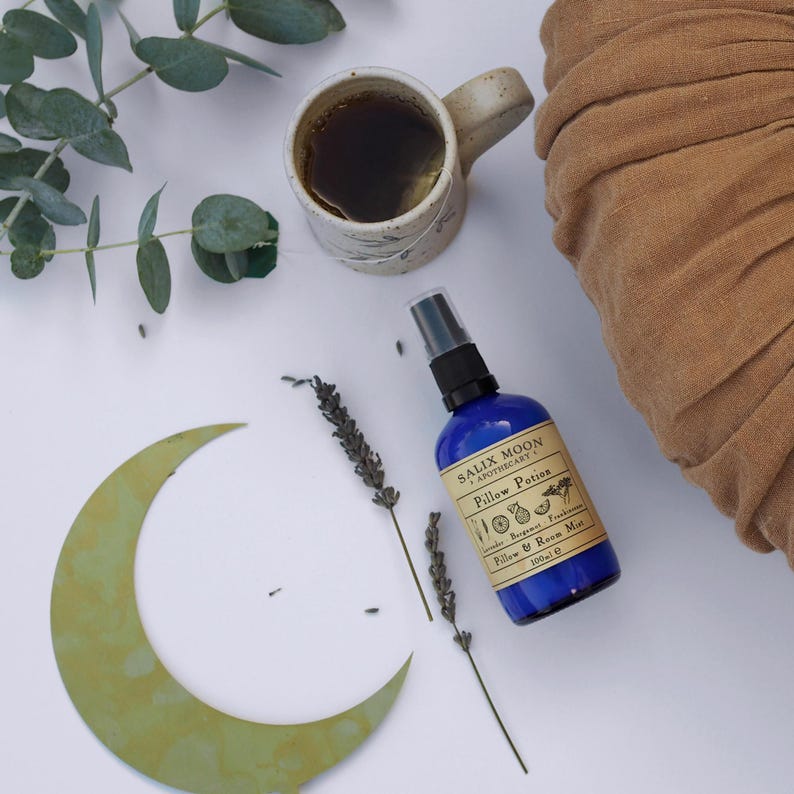 May include: A blue bottle of pillow and room mist with the text "Salix Moon Apothecary" and "Pillow Potion" on the label. The bottle is sitting on a white surface with a green crescent moon, lavender sprigs, and a brown ceramic mug with tea.