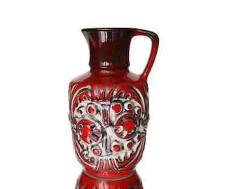 Puede incluir: Jarra de cerámica roja con tapa y asa marrón oscuro. El cuerpo presenta un diseño estampado en blanco y negro. La jarra es un artículo decorativo, probablemente para exhibición o servicio.